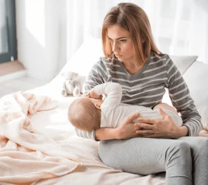 mom looking sad while breastfeeding baby while sitting on bed