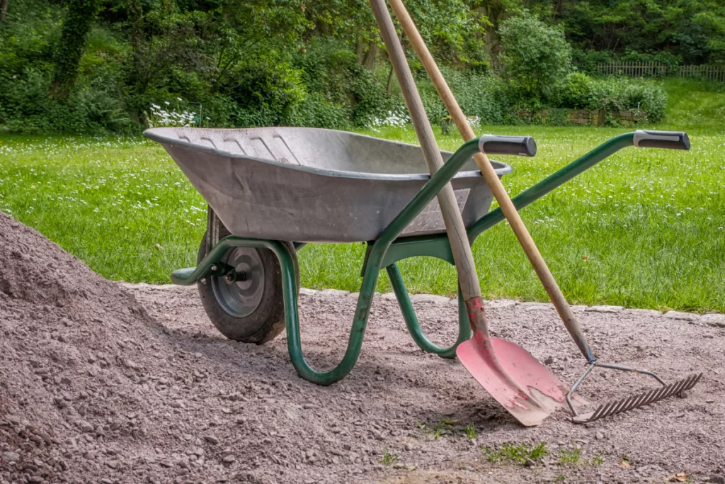 wheelbarrow with a shovel and garden rake leaning against it sitting next to a pile of garden soil