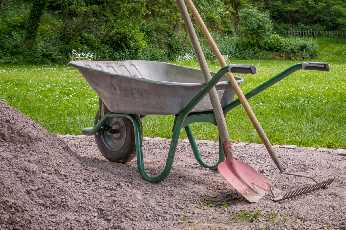 wheelbarrow next to a pile of compost with shovel and rake leaning up against it