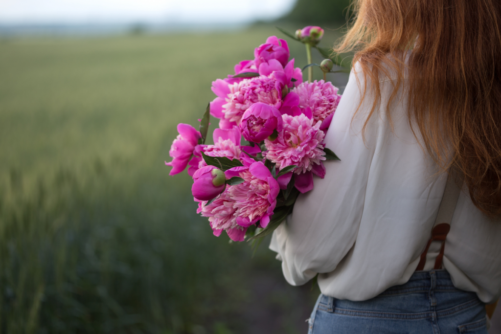 woman holding bouquet of peonies looking over green grain field