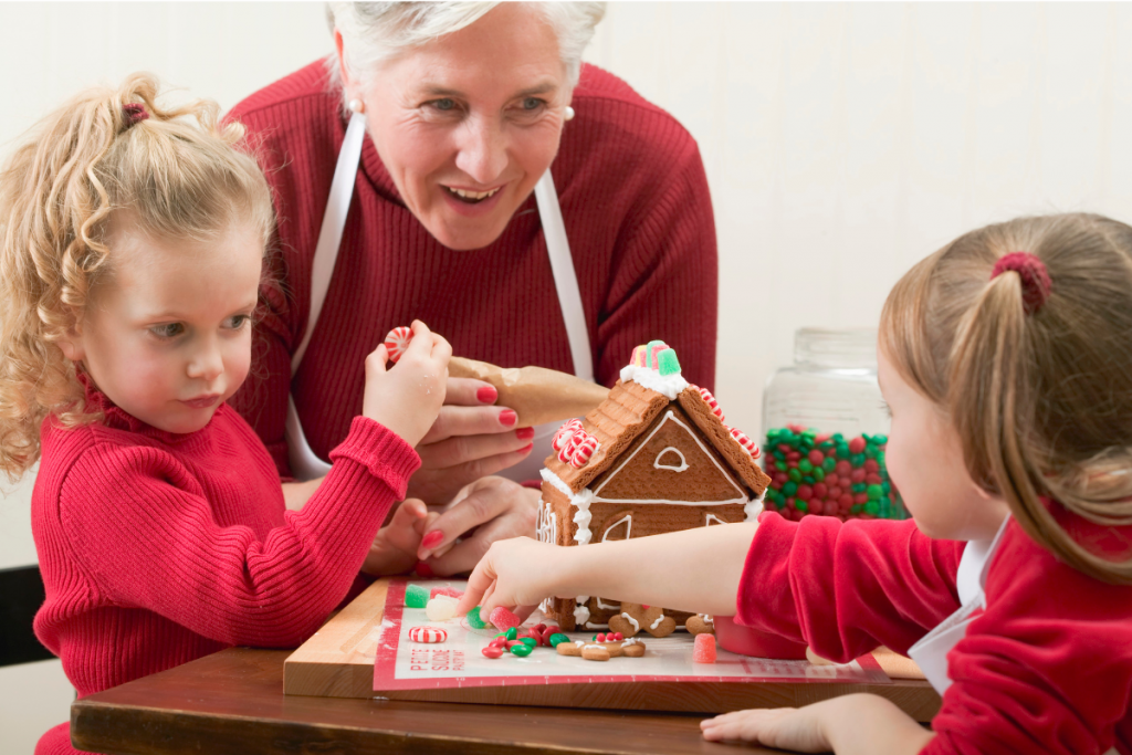 grandma and two granddaughters wearing red with white aprons doing a Christmas activity together by making a gingerbread house using green and red candy