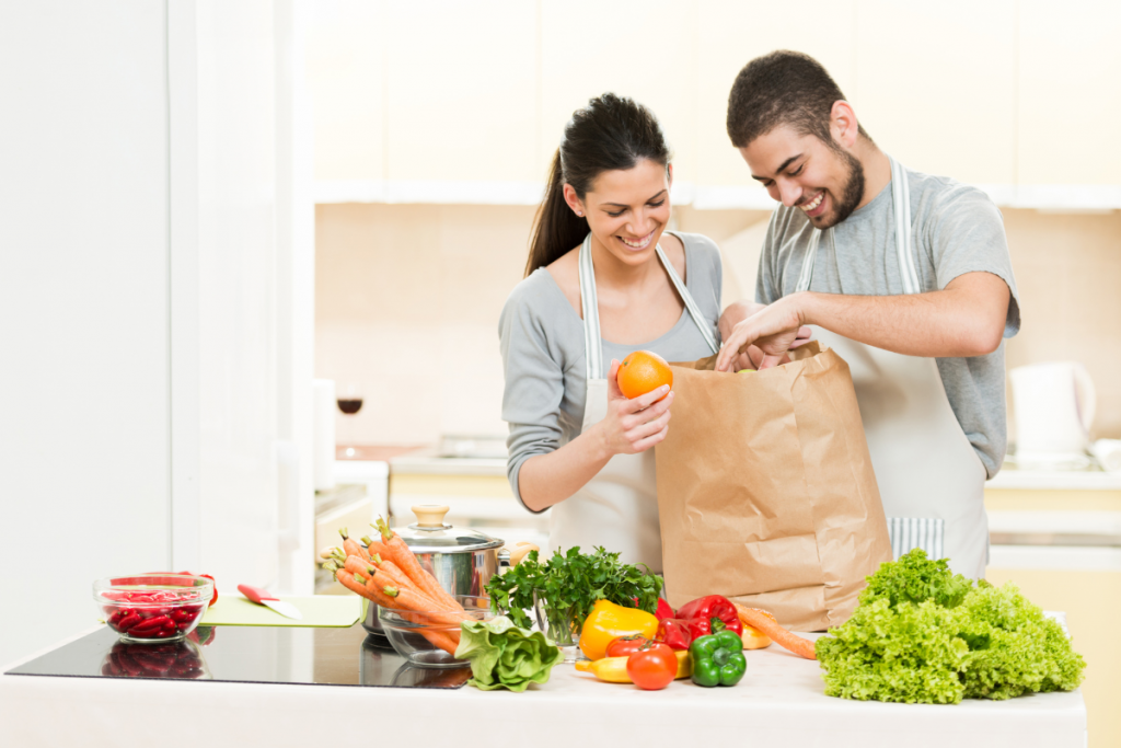 happy man and woman wearing aprons taking produce out of a brown paper grocery bag and setting them on the counter. counter is covered in fresh veggies and greens. 