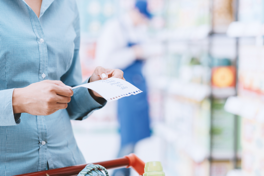 woman wearing a light blue shirt looking at her grocery shopping list standing in a grocery store next to the grocery cart