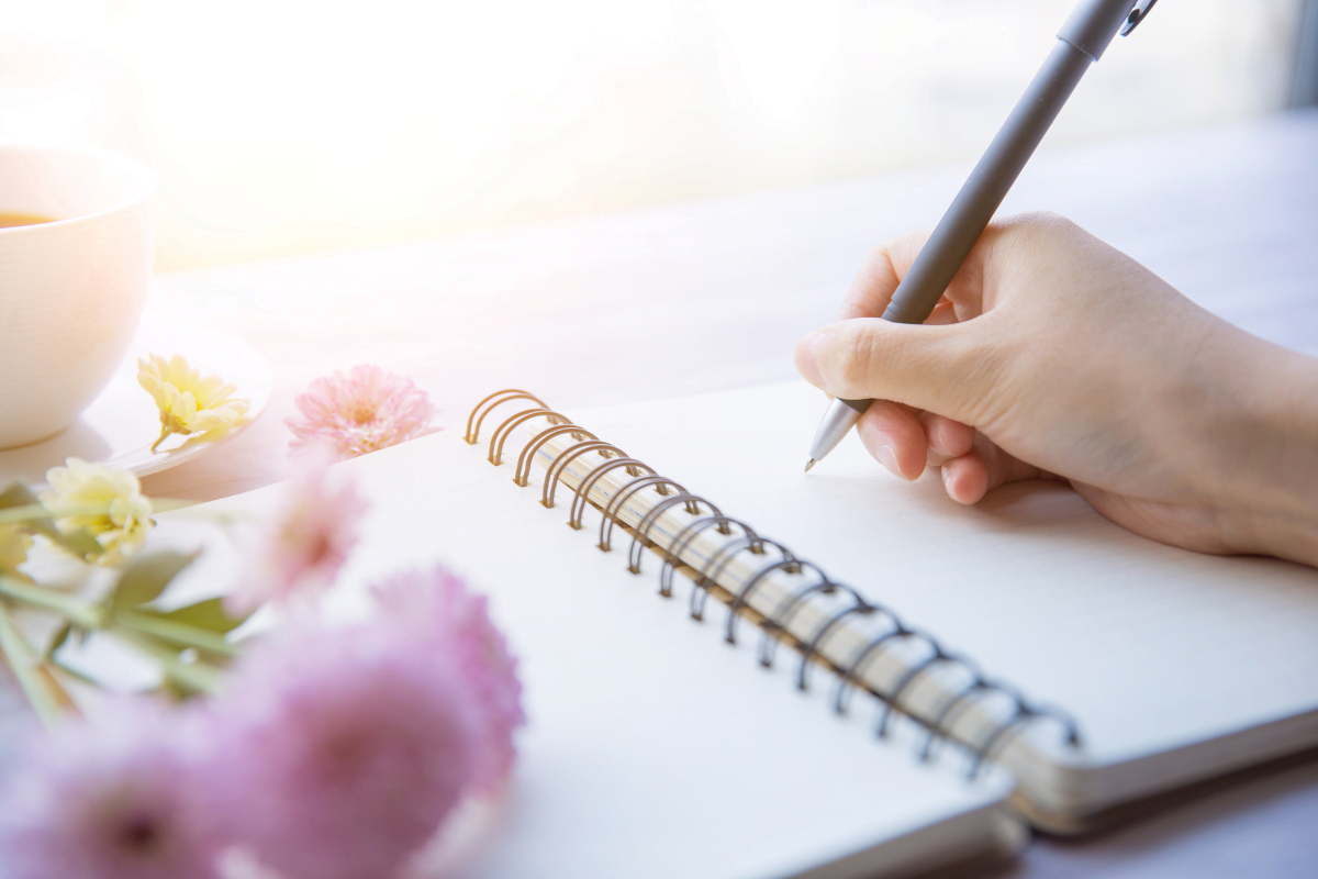 woman's hand holding grey pen writing on coiled notebook with a cub of tea and pink and white flowers beside the open notebook