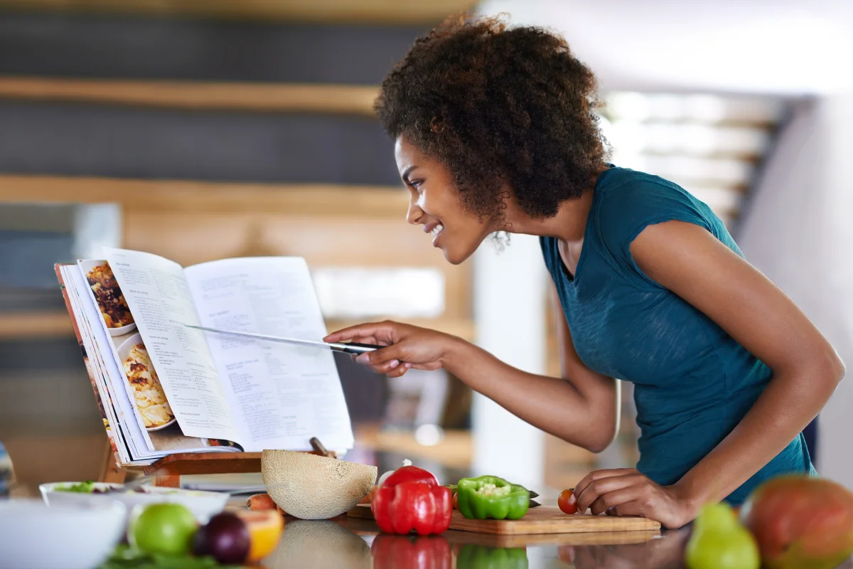 woman pointing at recipe book with knife while cutting fresh veggies on a wooden cutting board