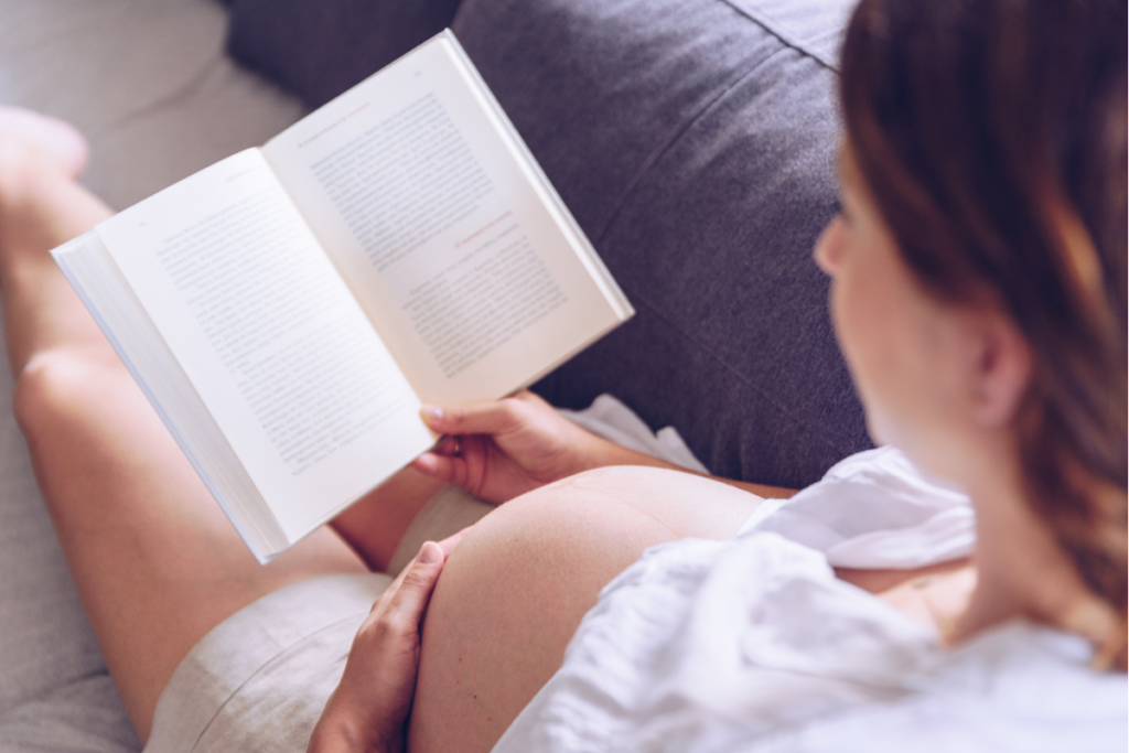 over the shoulder shot of a pregnant woman sitting on a couch and reading book holding naked pregnant belly