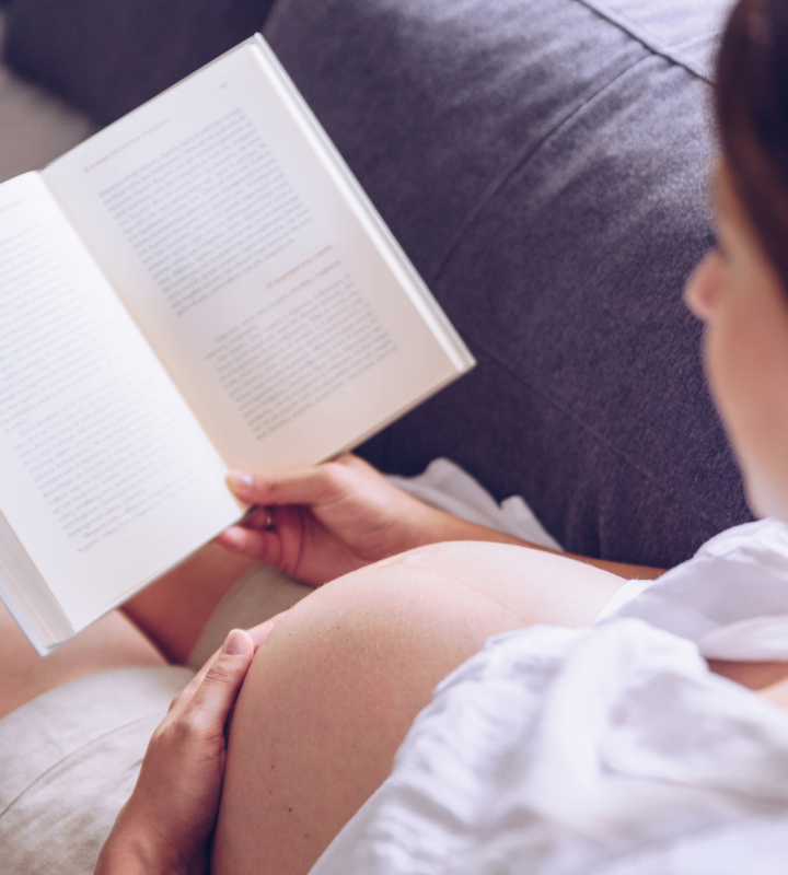 over the shoulder shot of a pregnant woman sitting on a couch and reading book holding naked belly