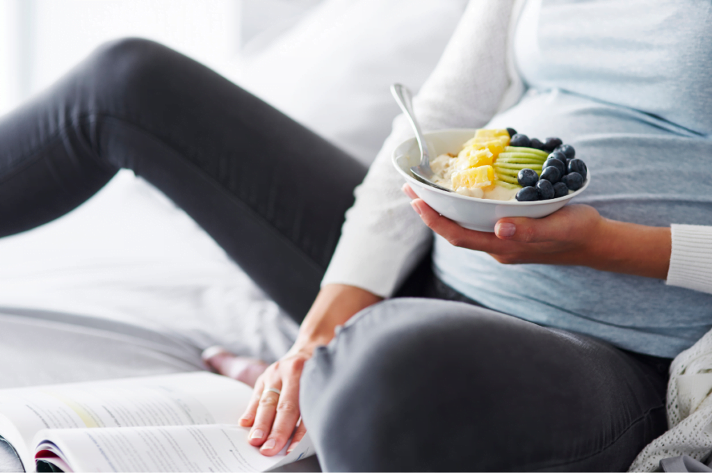 pregnant woman sitting on the floor wearing blue holding a bowl of fruit and yogurt while reading a book