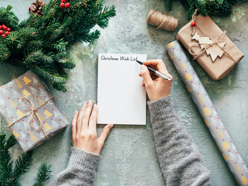 arms and hands, wearing grey sweater, writing on a notepad "Christmas Wish List" with black and grey pen. Surrounded by Christmas greenery, white gold and red Christmas Tree wrapping paper, brown wrapping paper with burlap twine. Roll of Christmas tree wrapping paper laying next to hands. 