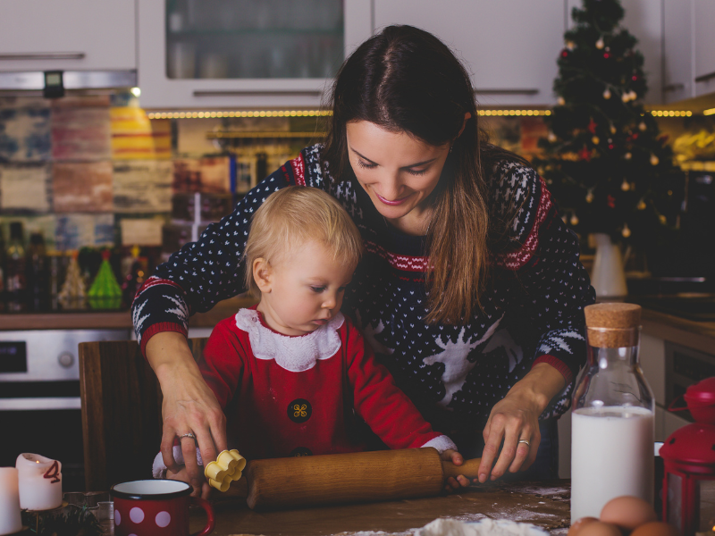 Mom helping toddler hold the rolling pin to bake. Woman is wearing Christmas sweater, Christmas tree on counter, toddler wearing red and white.
