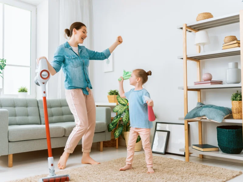 woman and girl dancing in living room with a vacuum and holding cleaning rag and spray bottle