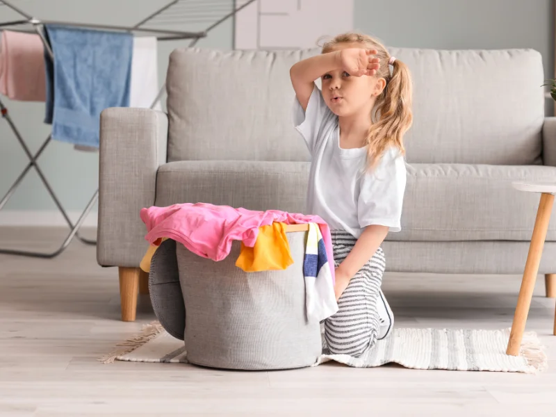 young girl looking tired sitting behind a basket of laundry in front of a couch