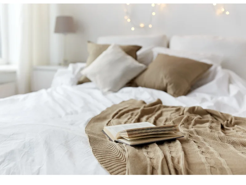 white bed, lights at head of bed, brown throw blanket and pillows with open book on top.
