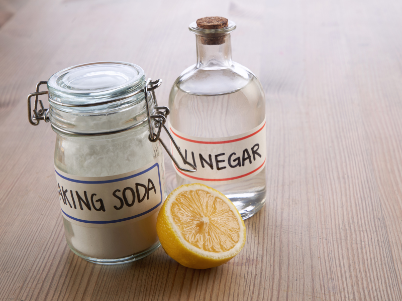 2 glass jars labeled vinegar and backing soda with a sliced lemon sitting in front it