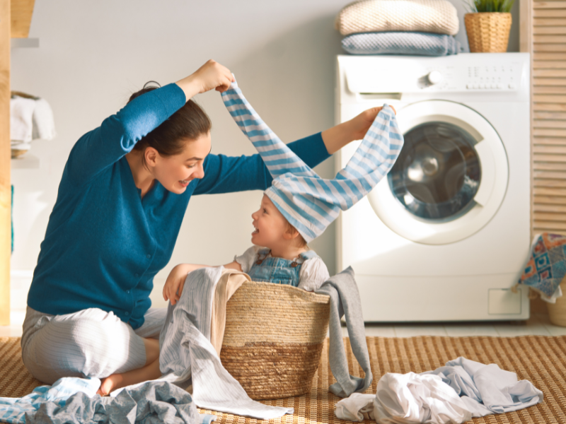 mom playing with young child in a laundry basked and laundry laying on floor in front of washing machine