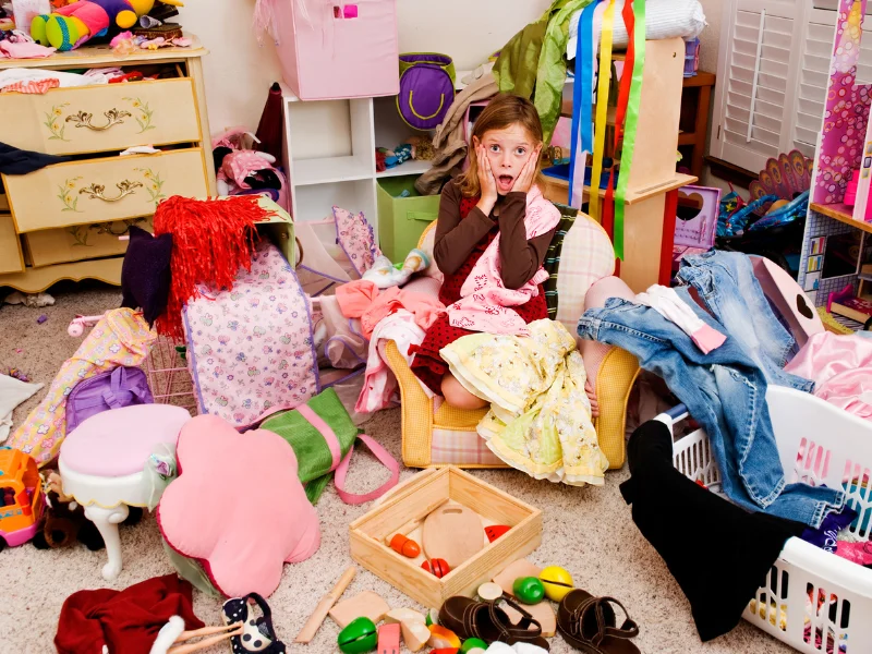 young girl sitting in the middle of a very messy room full of clothes and toys all over the floor