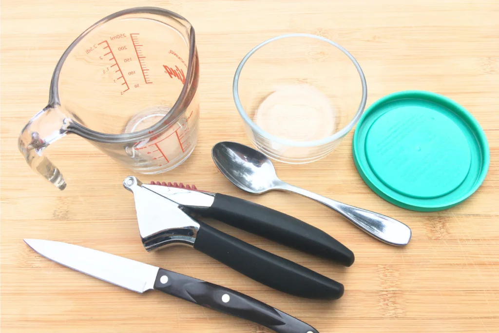 pyrex glass measuring 1 cup, glass pyrex glass bowl with teal lid, spoon, garlic press and Cutco paring knife sitting on a counter