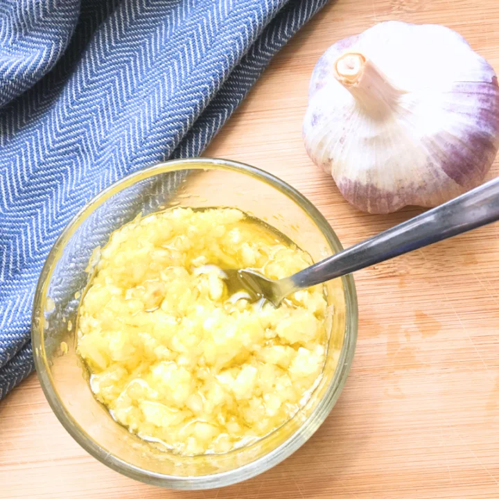 raw garlic olive oil spread in glass bowl next to a clove of garlic and a blue tea towel on a brown counter
