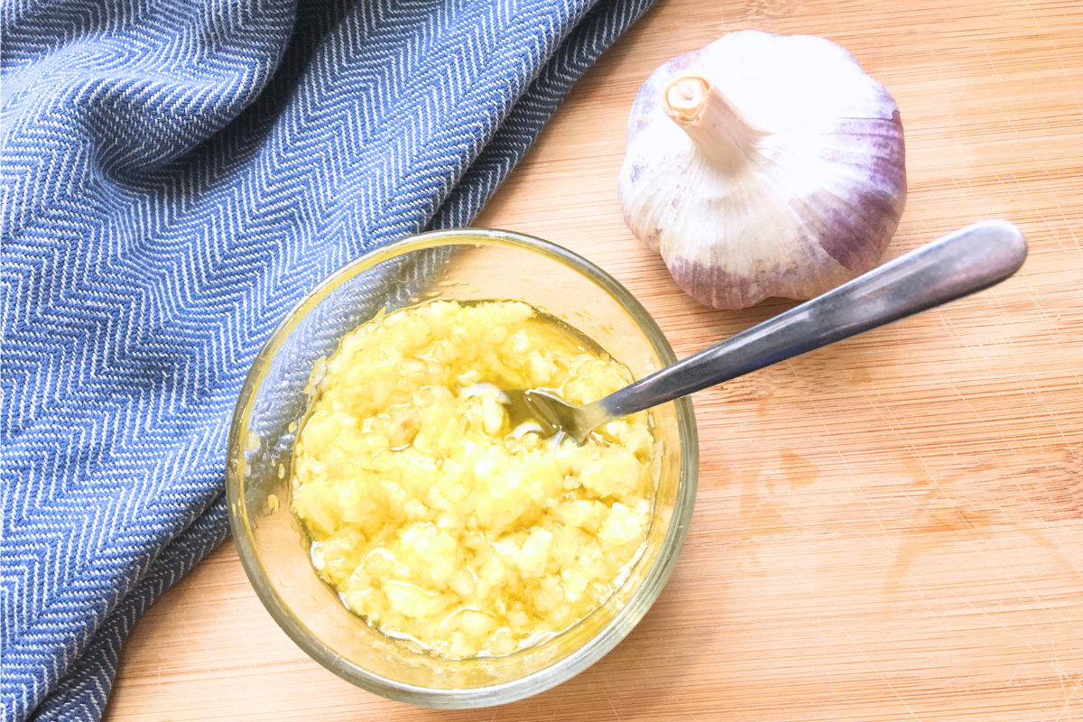 raw garlic olive oil spread in glass bowl next to a clove of garlic and a blue tea towel on a brown counter