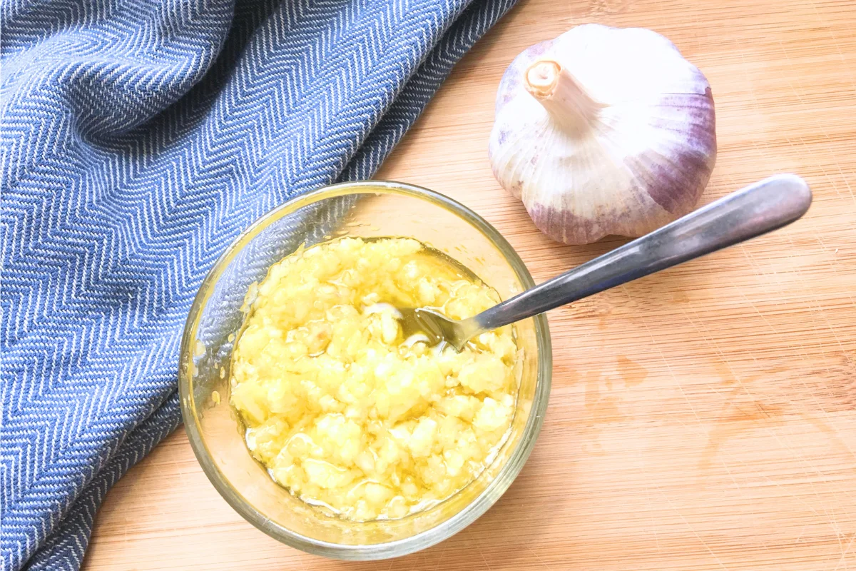 raw garlic olive oil spread in glass bowl next to a clove of garlic and a blue tea towel on a brown counter