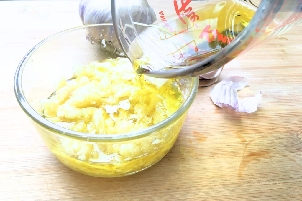 olive oil being poured overtop of crushed garlic in a glass bowl