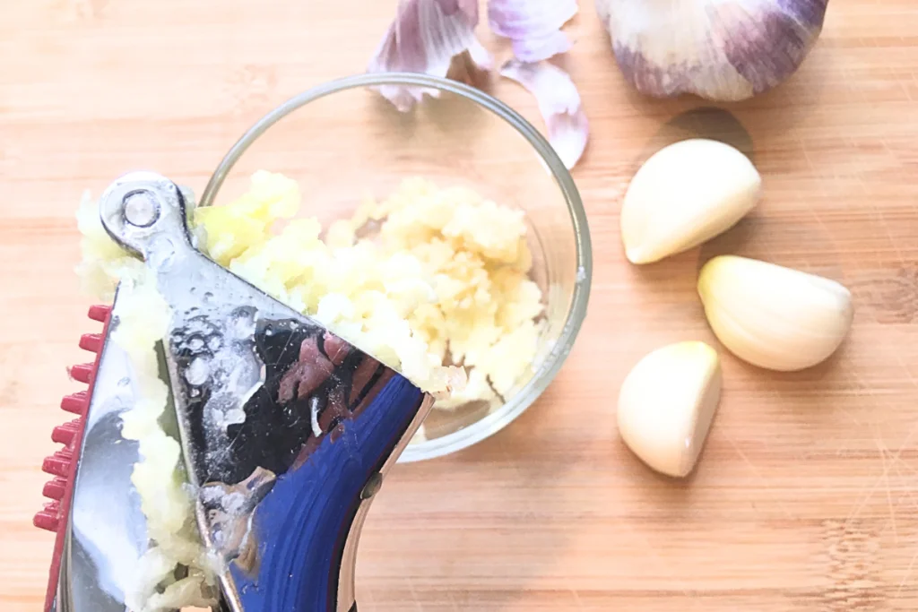 garlic being crushed in a garlic press into a glass bowl with peeled garlic cloves next to bowl