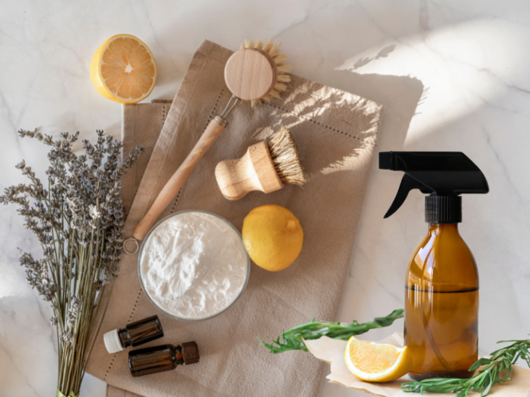essential oils, natural fibre wooden scrub brushes, lemons, white powder, amber spray bottle, lavender on brown towel on white marble surface.