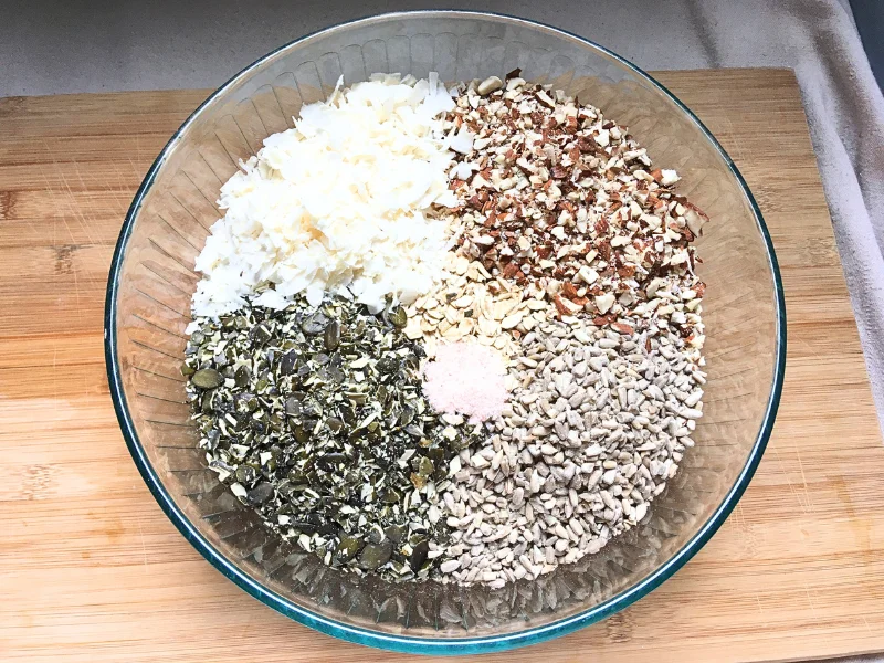 glass bowl containing these ingredients: coconut flakes, chopped almonds, sunflower seeds, chopped pumpkin seeds, salt, rolled oats