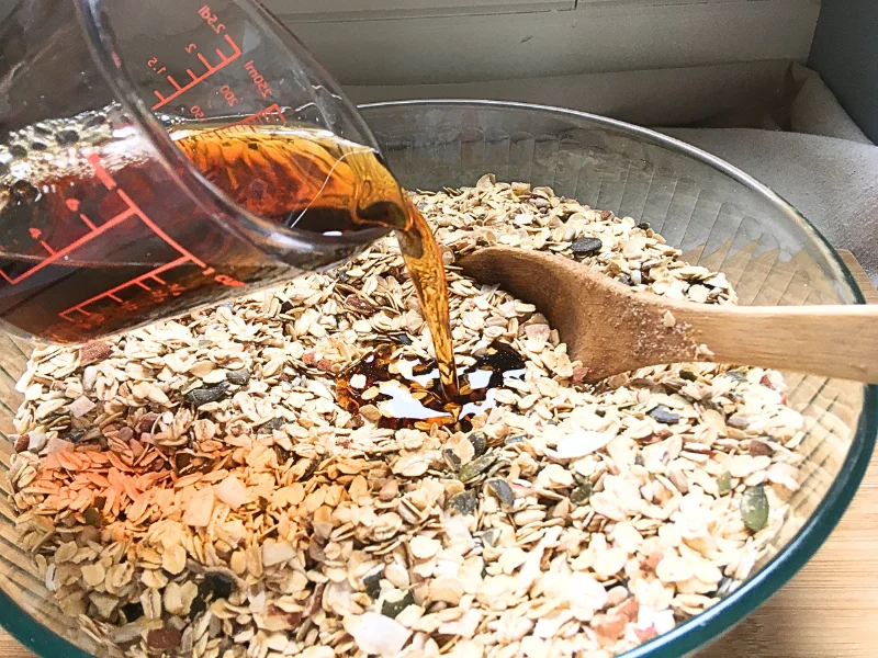 pouring maple syrup into the dry granola ingredients with wooden spoon in bowl