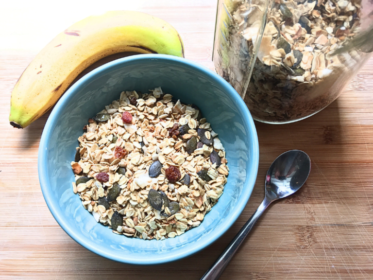 healthy granola in teal bowl with banana and spoon next to it. Mason jar with granola also next to it.
