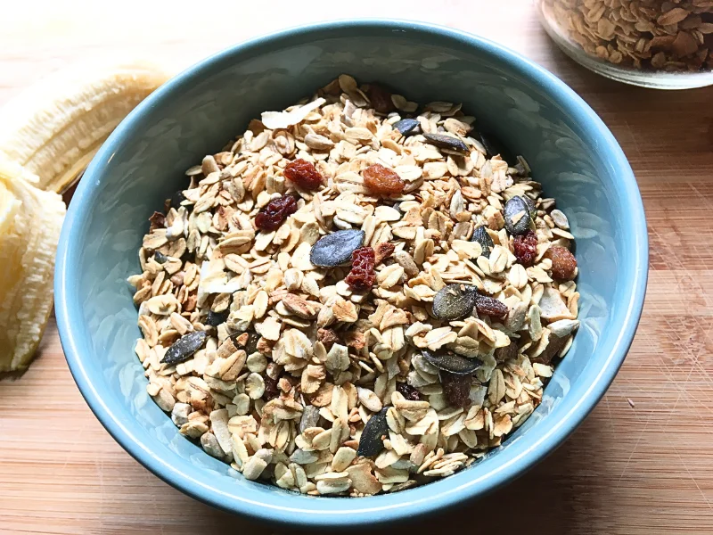 homemade healthy granola with raisins in teal bowl with banana next to it.