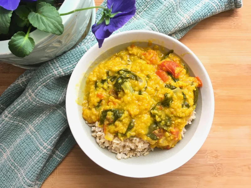 red lentil coconut curry served on top of brown rice in a bowl on a wooden counter top with purple pansies and a teal towel in the background.