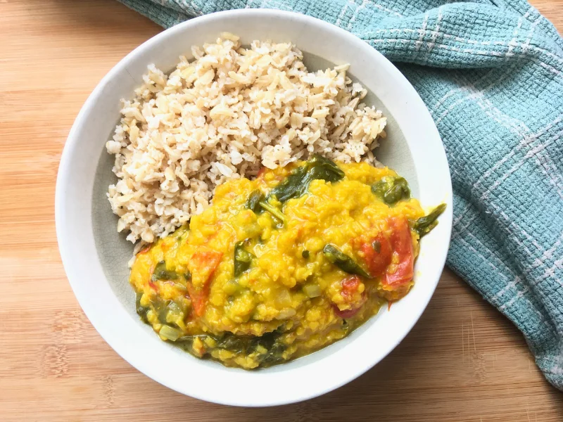 red lentil coconut curry with greens and tomatoes next to brown rice in a bowl on a wooden counter with a tea towel
