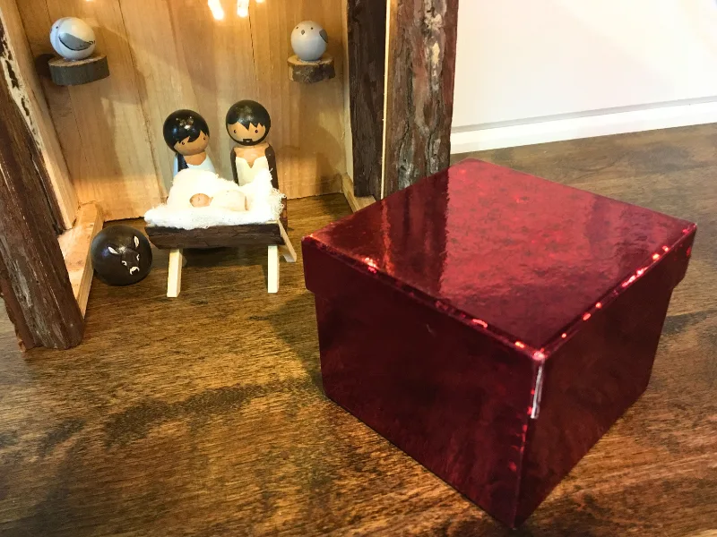 Painted wooden peg nativity scene with baby Jesus in the manger, Mary & Joseph in the stable and a red Christmas box in foreground.