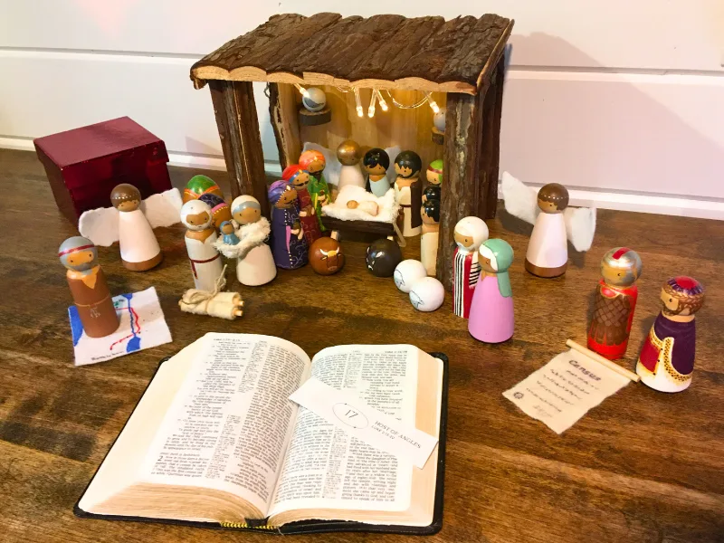 Painted wooden peg nativity scene with Marry, Joseph, Baby Jesus, donkey, sheep, camels, magi, and shepherds near the stable. Red Christmas box and Bible.