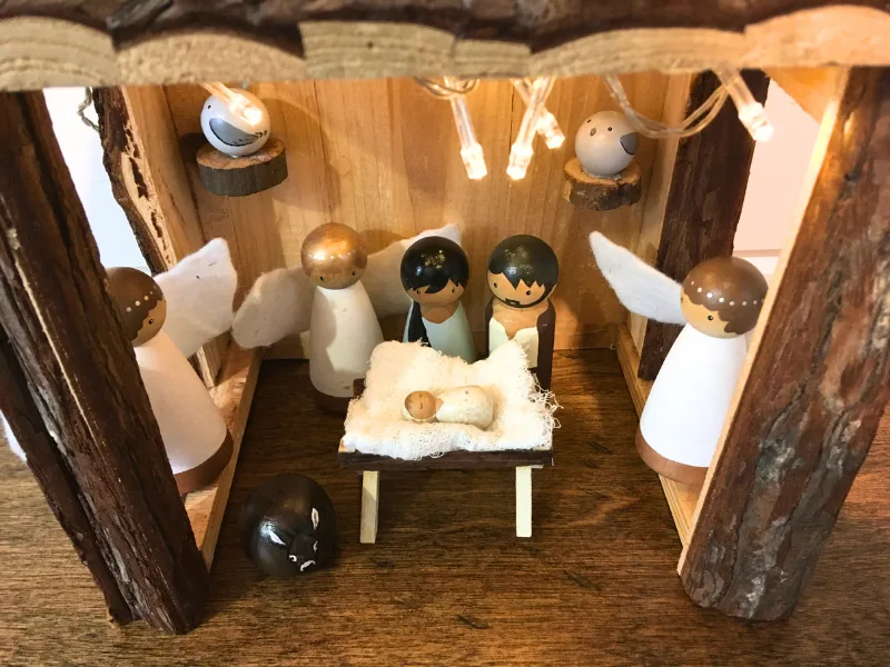 Wooden peg nativity scene with Marry, Joseph, Baby Jesus, donkey, doves, and angels in a stable with lights.