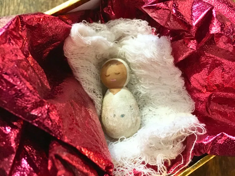 Wooden peg baby Jesus in swaddling cloth, on red shiny tissue paper in a gold box.