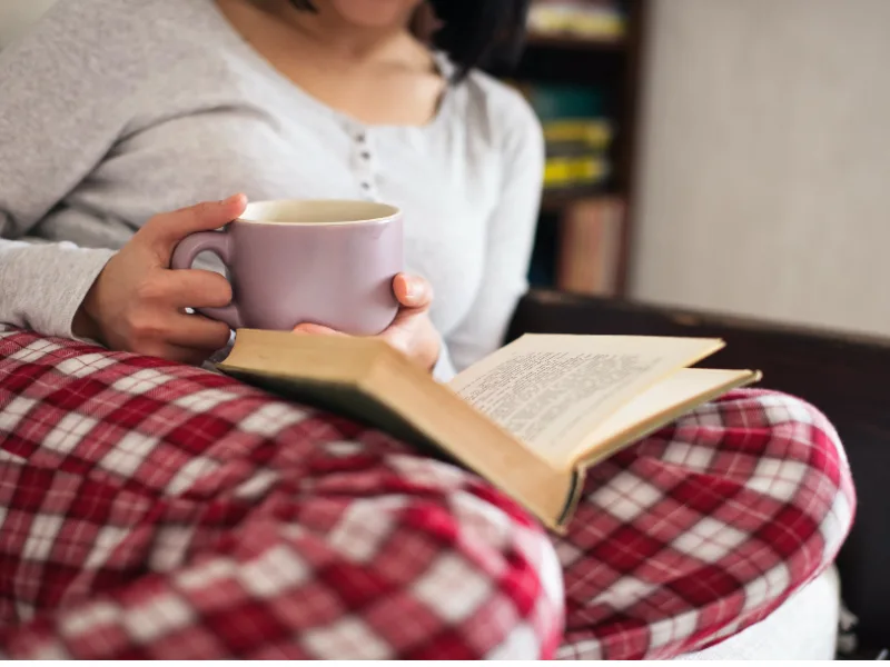 woman wearing red and white plaid pants holding a mug and reading a book