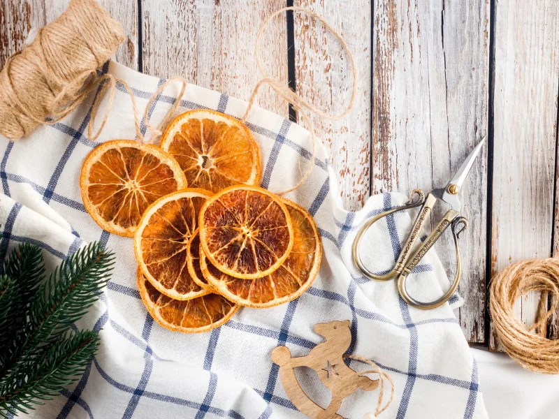 diy homemade dried oranges on white tea towel on wooden table with burlap strings, scissors, wooden rocking horse and spruce greenery.