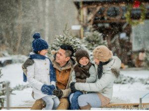 family of four bundled up and sitting outside by a cabin and spruce trees