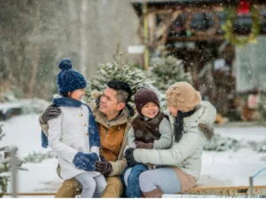 family of four bundled up and sitting outside by a cabin and spruce trees
