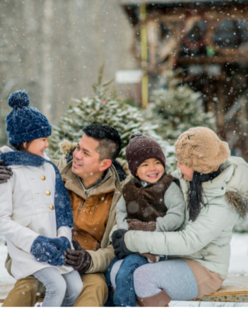family of four bundled up and sitting outside by a cabin and spruce trees