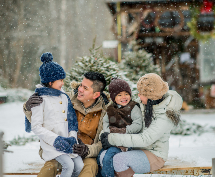 family of four bundled up and sitting outside by a cabin and spruce trees