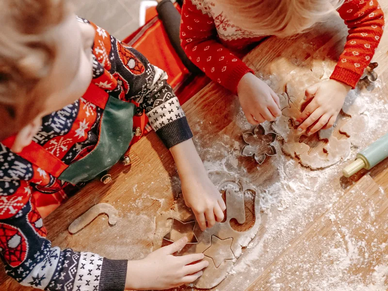 toddlers dressed in Christmas sweaters cutting out cookie shapes on floured table