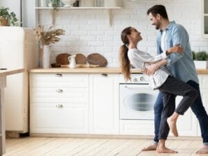 couple dancing in minimalist clean decluttered kitchen with wooden counter tops, white cupboards, wooden cutting boards plants and open shelving