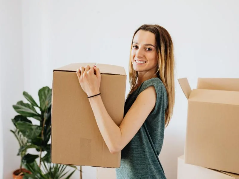 smiling woman holding cardboard box with another one behind and plants in the corner