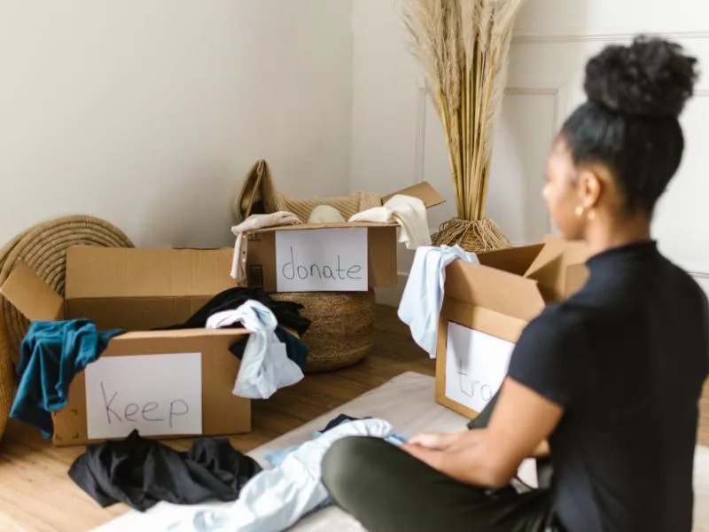 woman sitting on floor looking at donation boxes with stuff in them and clothes on the floor