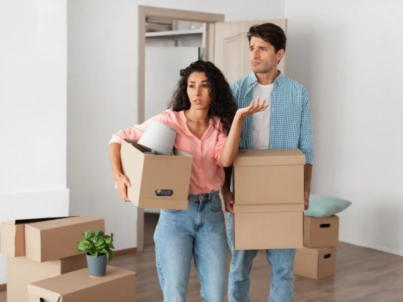 man and woman looking confused holding cardboard boxes surrounded by more cardboard boxes