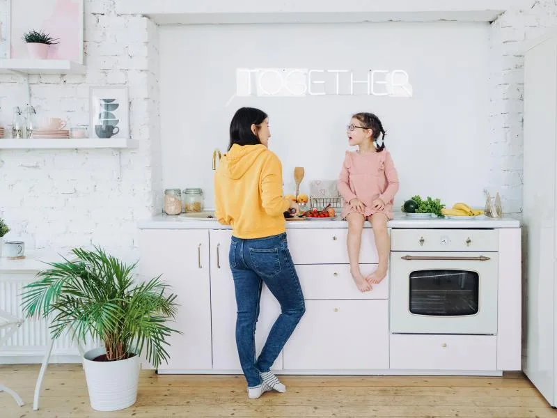 mom cooking food in tidy white kitchen while daughter sits on counter and talks to her