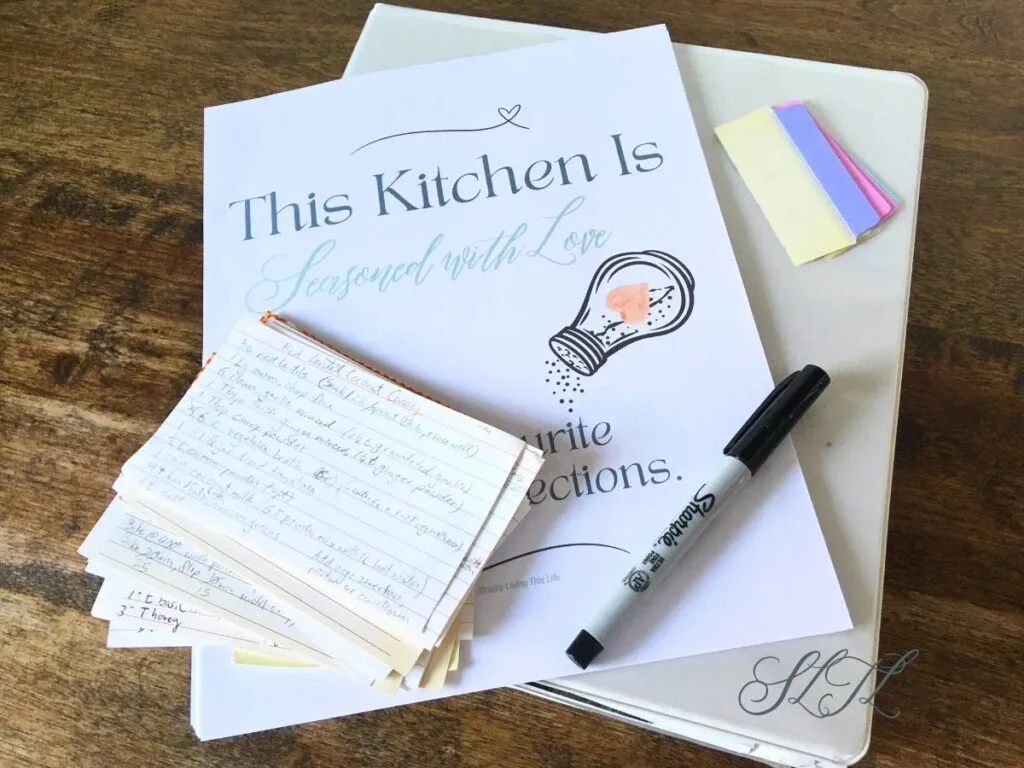white binder on wooden counter with recipe binder ebook printable, black sharpie, binder dividers and stack of recipe cards